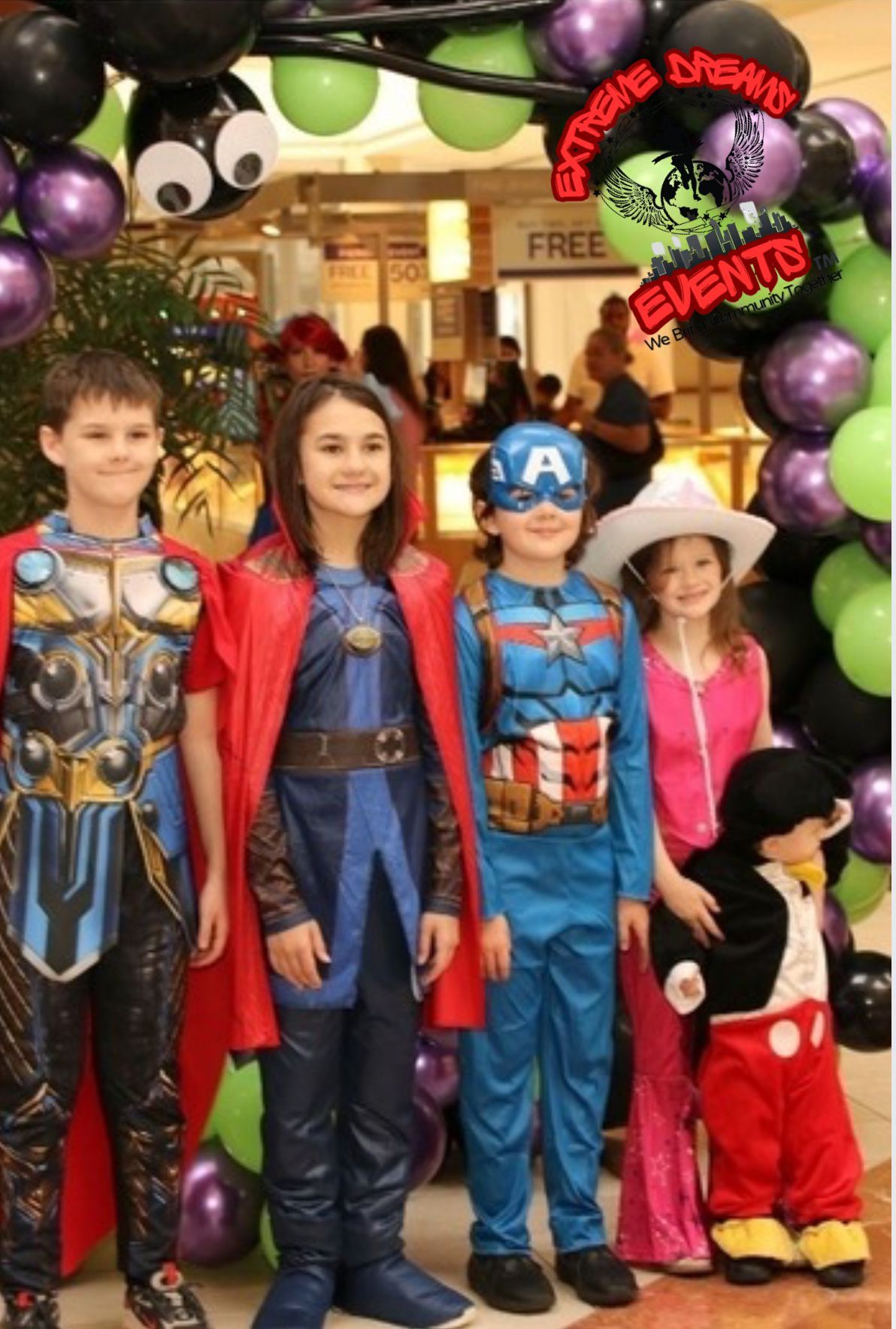 Four children in superhero costumes stand under a balloon arch at a store entrance