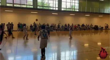 Indoor basketball game in a gym, with players on court and spectators seated along the wall