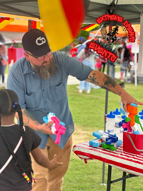 Vendor handing toys at an outdoor event booth with colorful toys on a red table