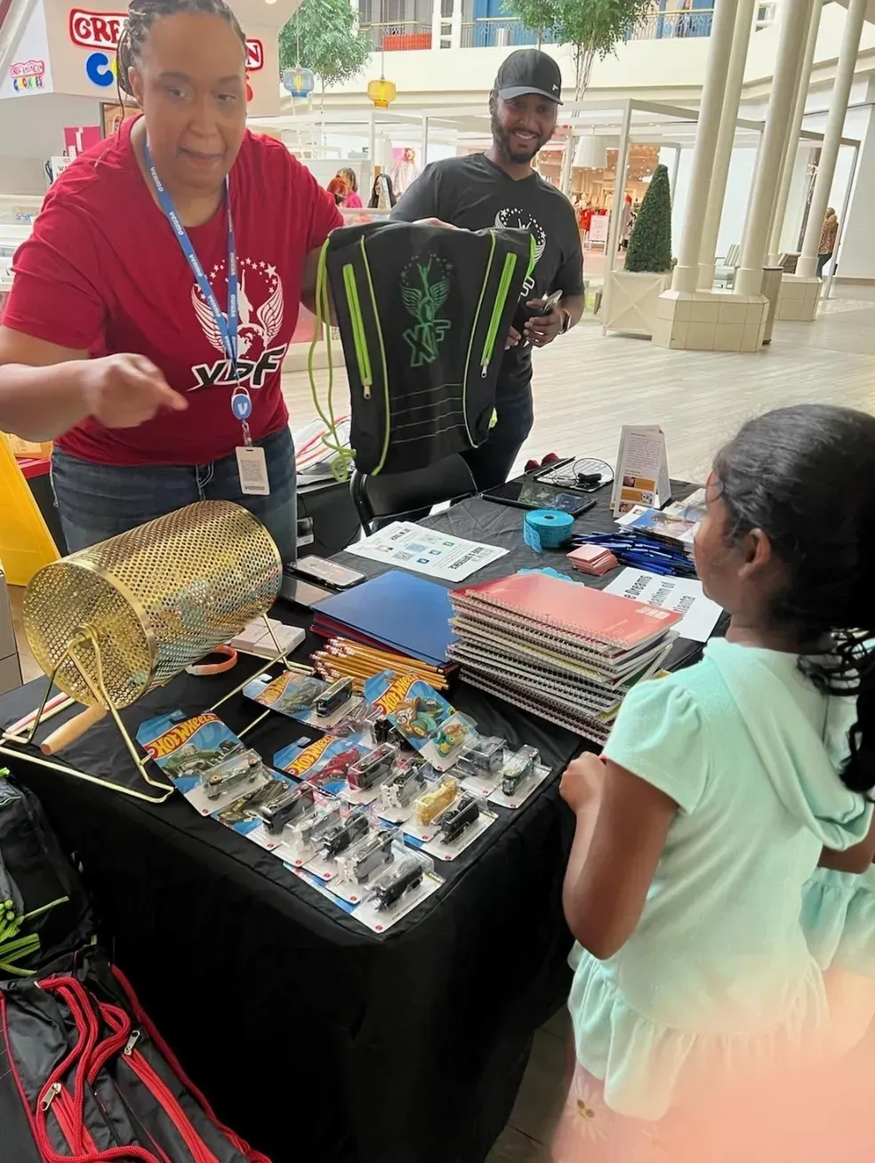 Vendor booth at a mall event displaying collectible action figures and merchandise with people interacting.
