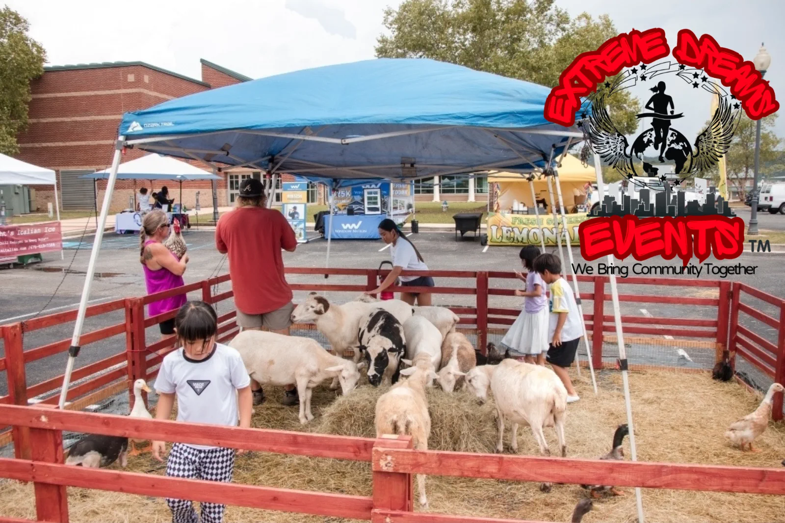 Children petting goats in a red-fenced pen at an outdoor farm event under a blue canopy